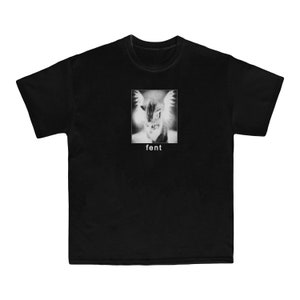Puede incluir: Camiseta negra con un gráfico de un gato con alas. El texto "fent" está impreso debajo del gráfico.
