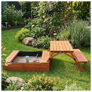 May include: A wooden sandbox with a black liner and a matching picnic table set on a green lawn. The sandbox contains sand and small toys. The picnic table has a folding design.