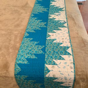 May include: A quilted bed runner in shades of turquoise and white. The runner features a central band of solid turquoise, flanked by patterned sections with a repeating arrow design. The white sections have a dragonfly and floral pattern.