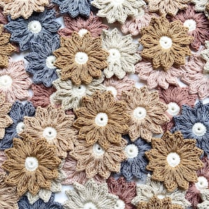 May include: A collection of handmade crochet flowers in various neutral colors, including beige, brown, pink, and blue. Each flower has a white center and detailed petal design, perfect for crafting projects.