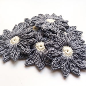 May include: A group of gray crocheted flowers with white centers. The flowers are arranged in a pile on a white background.