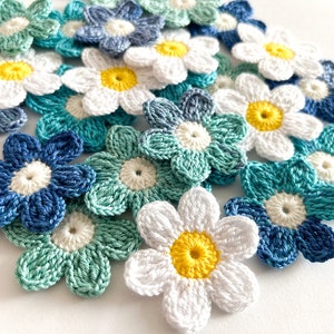 Puede incluir: Una colección de flores de ganchillo en blanco, azul y verde. Las flores tienen un diseño sencillo con cinco pétalos y un círculo central.