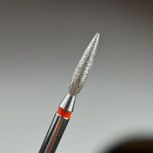 May include: A close-up of a silver diamond nail drill bit with a red band. The bit is pointed and has a rough texture.