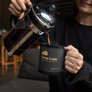 May include: A black coffee mug with the text "Error Code" and a cloud graphic is being filled with coffee from a French press. The mug also has the text "Find refreshment with coffee or tea."
