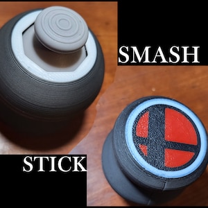 Op de afbeelding: Twee 3D-geprinte zwarte en witte joystick-kappen. Eén kap heeft een witte ring met een grijze knop in het midden. De andere kap heeft een witte ring met een rood en zwart Smash-logo in het midden.