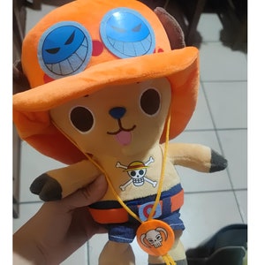 May include: A plush toy of Tony Tony Chopper from the anime One Piece. Chopper is wearing his signature orange hat with blue skull and crossbones markings, a blue and orange belt, and a necklace with a skull pendant.