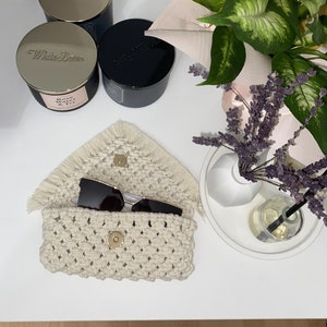 May include: A white crocheted bag with a fringe border and a gold button closure. The bag is open and sunglasses are inside. The bag is on a white surface with candles and a plant in the background.