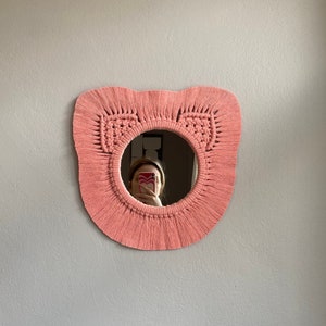 May include: A pink macrame mirror with a round mirror in the center. The mirror is hanging on a white wall.