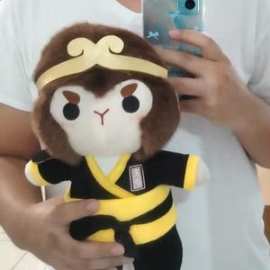May include: Plush toy of a monkey character with a brown head, gold headband, and a black and yellow outfit. The monkey has a white face, black eyes, and a red mouth. The toy is held in front of a white shirt.