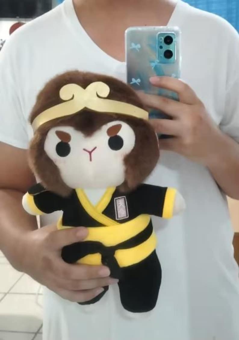 May include: Plush toy of a monkey character with a brown head, gold headband, and a black and yellow outfit. The monkey has a white face, black eyes, and a red mouth. The toy is held in front of a white shirt.