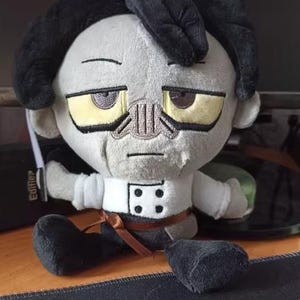 Può includere: Una bambola di peluche raffigurante un personaggio dalla pelle grigia con capelli neri, occhi gialli e un dettaglio del naso cucito. Il personaggio indossa una camicia bianca con quattro bottoni neri e una cintura a nastro marrone, con pantaloni neri.