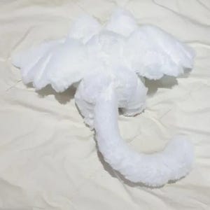 Puede incluir: Juguete de drag&oacute;n de peluche blanco con alas y una cola larga. El drag&oacute;n est&aacute; acostado de espaldas con las alas extendidas.