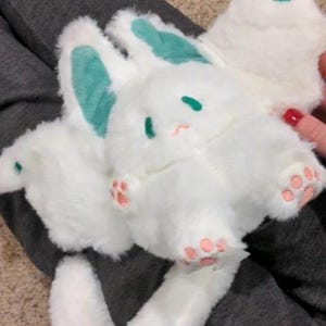 Puede incluir: Juguete de peluche blanco con una cola larga, orejas verdes y huellas de patas rosas. El juguete tiene un cuerpo redondo y una cara linda de dibujos animados.