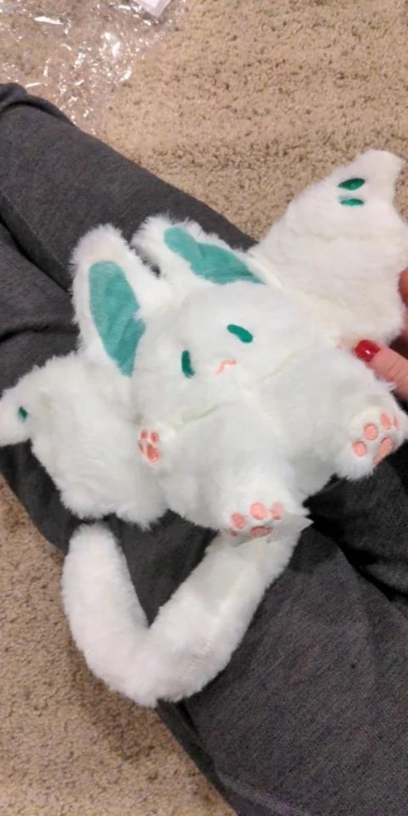 Puede incluir: Juguete de peluche blanco con una cola larga, orejas verdes y huellas de patas rosas. El juguete tiene un cuerpo redondo y una cara linda de dibujos animados.