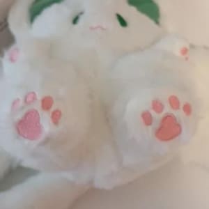 Puede incluir: Conejo de peluche blanco con orejas verdes y huellas de patas rosas. El conejo est&aacute; acostado de lado con la cabeza girada.