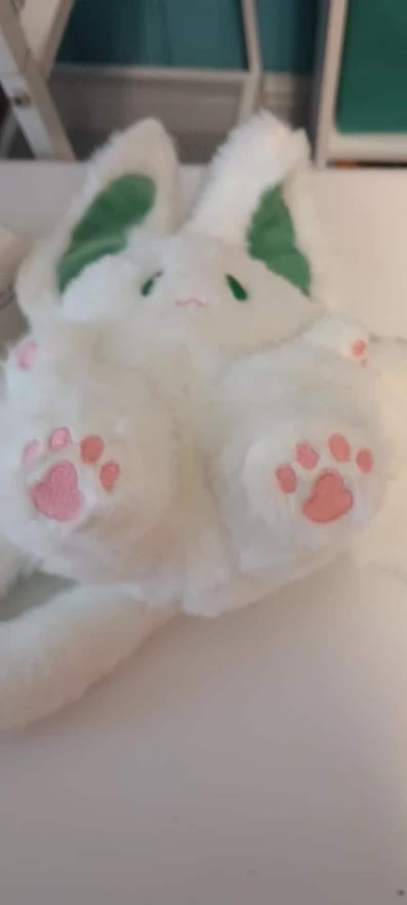 Puede incluir: Conejo de peluche blanco con orejas verdes y huellas de patas rosas. El conejo est&aacute; acostado de lado con la cabeza girada.