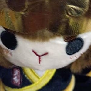 May include: A plush toy featuring a monkey-like character with a brown fur head and a gold-colored crown. The character has black eyes, a white face, and a red embroidered nose. It wears a black and yellow outfit.