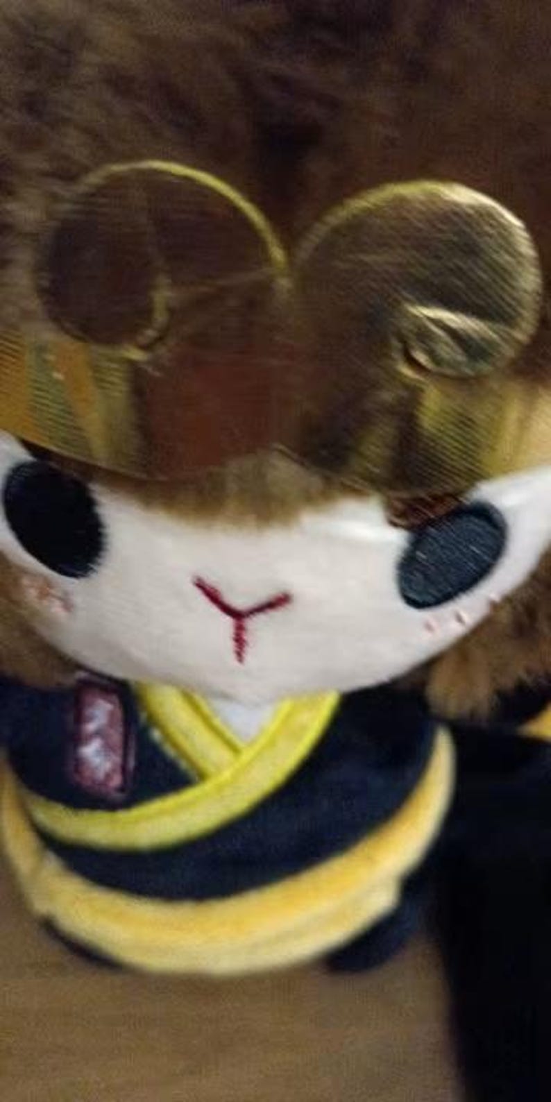 May include: A plush toy featuring a monkey-like character with a brown fur head and a gold-colored crown. The character has black eyes, a white face, and a red embroidered nose. It wears a black and yellow outfit.
