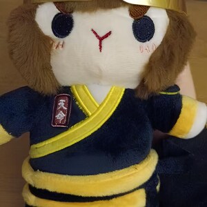 May include: Plush toy of a monkey-like character wearing a gold headband and a dark blue outfit with yellow trim. The toy has a white face with embroidered eyes and a red mark. The outfit has a yellow sash and a small patch with Chinese characters.
