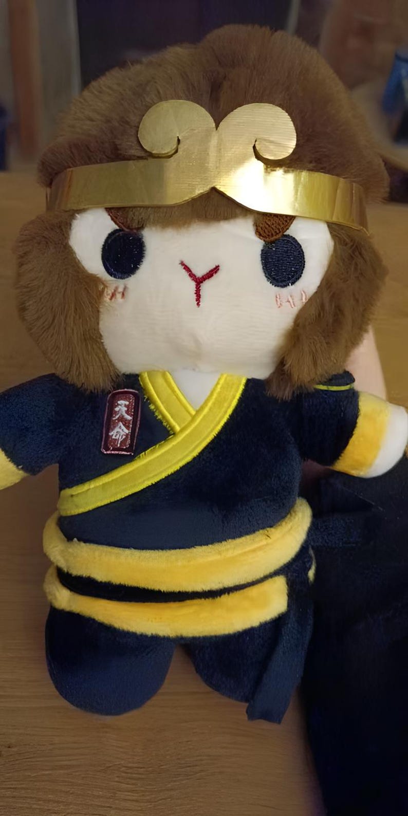 May include: Plush toy of a monkey-like character wearing a gold headband and a dark blue outfit with yellow trim. The toy has a white face with embroidered eyes and a red mark. The outfit has a yellow sash and a small patch with Chinese characters.