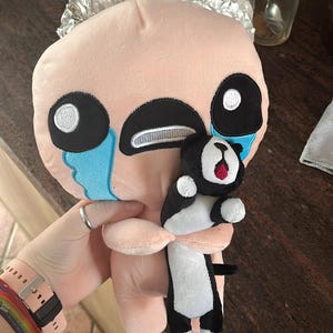 May include: A pink plush toy with a black face and blue teardrop-shaped eyes. The toy is holding a smaller black and white plush toy with a red tongue sticking out. The larger toy has the text "bomilk" on its chest.