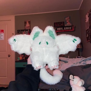 Puede incluir: Juguete de peluche blanco con orejas verdes y una cola larga. El juguete tiene una cara linda y de dibujos animados con ojos grandes. El juguete est&aacute; siendo sostenido en la mano de una persona.