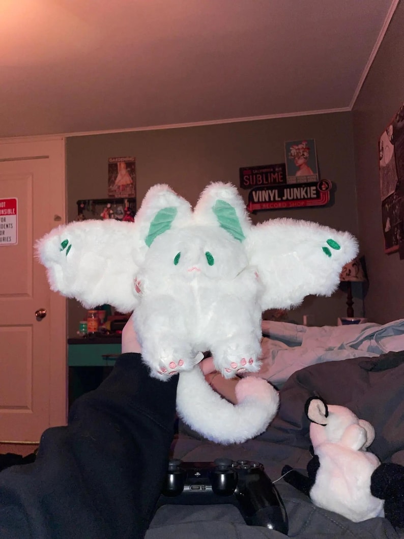 Puede incluir: Juguete de peluche blanco con orejas verdes y una cola larga. El juguete tiene una cara linda y de dibujos animados con ojos grandes. El juguete est&aacute; siendo sostenido en la mano de una persona.