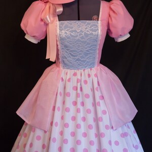 May include: A pink and white polka dot dress with a matching pink bonnet. The dress has a blue lace bodice and a pink sash.