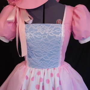 May include: A pink dress with a white and pink polka dot skirt, a blue lace bodice, and a matching pink bonnet with a ribbon.