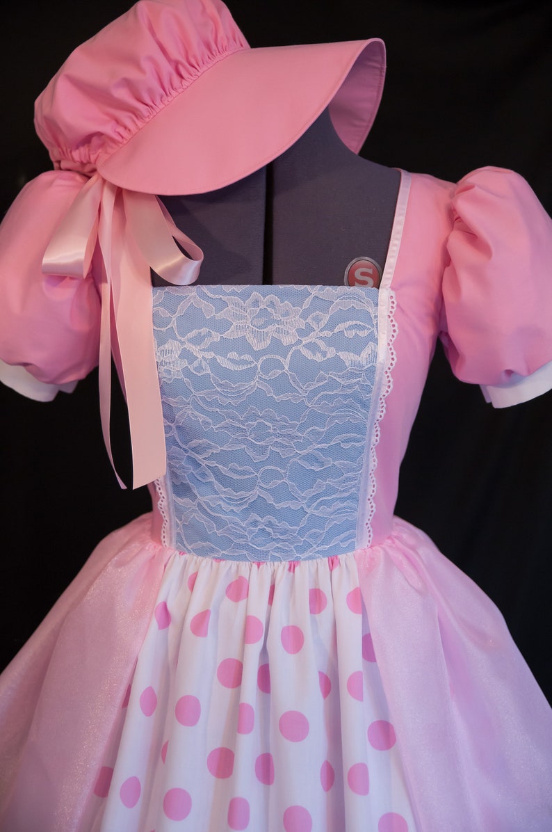 May include: A pink dress with a white and pink polka dot skirt, a blue lace bodice, and a matching pink bonnet with a ribbon.