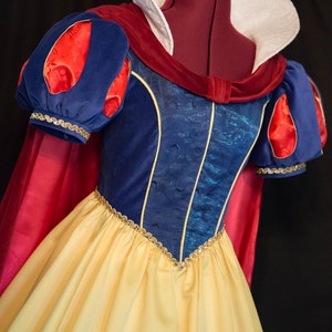 May include: A blue and yellow Snow White costume with a red cape and white collar. The costume features a blue bodice with a gold trim and a yellow skirt.