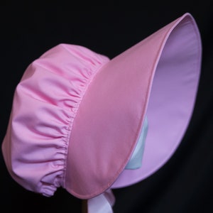 May include: A pink sunbonnet with a gathered crown and a wide brim. The bonnet is made of a soft, lightweight fabric.