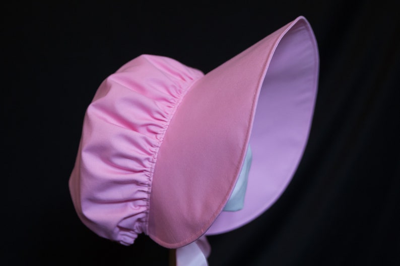 May include: A pink sunbonnet with a gathered crown and a wide brim. The bonnet is made of a soft, lightweight fabric.