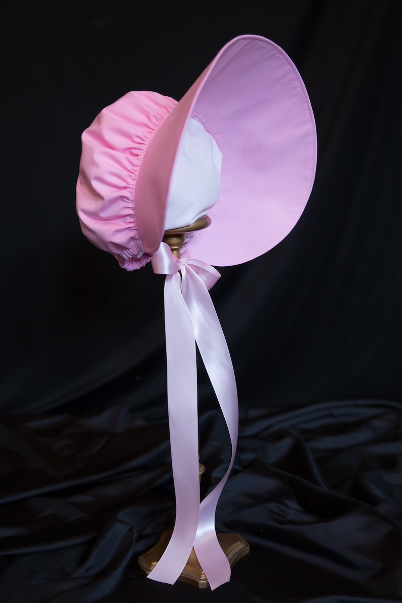 May include: A pink bonnet with a large brim and a white lining. The bonnet is tied with a pink ribbon and is displayed on a gold stand.