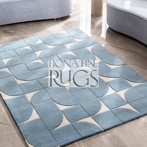 Puede incluir: Una alfombra rectangular con un patrón geométrico de formas azul claro y blanco roto. La alfombra presenta un diseño repetitivo de elementos de cuarto de círculo y cuadrados. El texto "FIONA FINE RUGS" está centrado en la alfombra.
