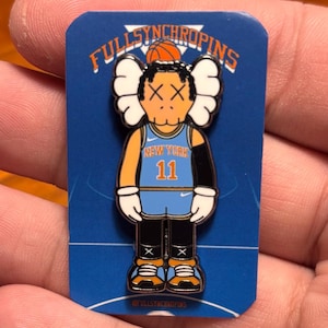 May include: A pin featuring a cartoon character in a New York Knicks jersey with the number 11. The pin is on a blue backing with the words "FULLSYNCHROPINS" at the top. The character has white eyes and a black outline.