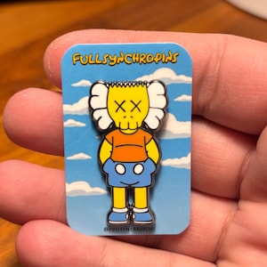 May include: A rectangular pin featuring a cartoon character with an orange head, yellow body, blue shorts, and blue shoes. The character has white ears and crossed-out eyes. The pin has the text "FULLSYNCHROPINS" on a blue background with white clouds.
