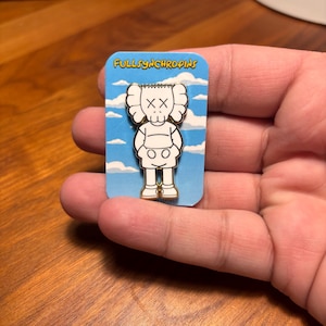 Pode incluir: Um alfinete de esmalte branco e preto com um personagem de desenho animado com olhos riscados e um corpo arredondado. O alfinete est&aacute; em um cart&atilde;o azul com nuvens brancas e o texto "FULLSYNCHROPINS".