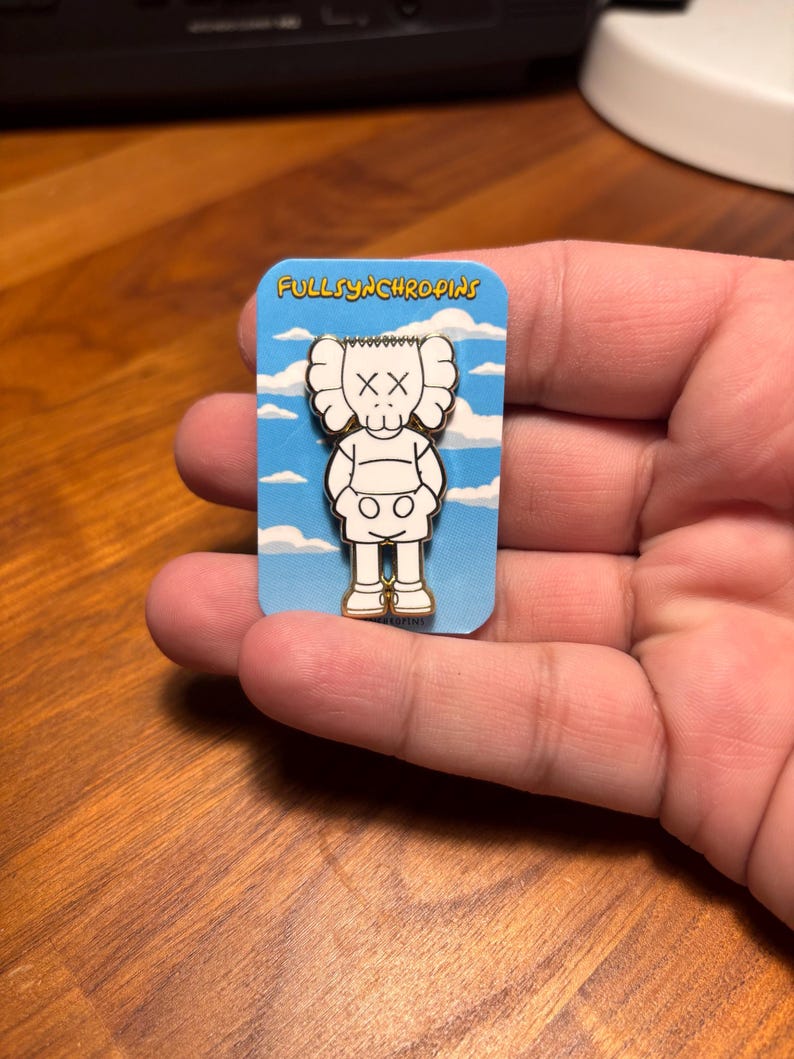 Pode incluir: Um alfinete de esmalte branco e preto com um personagem de desenho animado com olhos riscados e um corpo arredondado. O alfinete est&aacute; em um cart&atilde;o azul com nuvens brancas e o texto "FULLSYNCHROPINS".