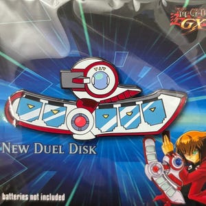 Puede incluir: Un pin de esmalte rojo, blanco y azul con forma de Disco de Duelo de Yu-Gi-Oh! GX. El pin presenta el texto "New Duel Disk" y está empaquetado sobre un fondo azul con una ilustración de personaje.