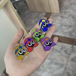 May include: A collection of colorful enamel pins featuring a cartoon character with a round black body, a curved white mouth, and a hat with various color schemes. The pins are yellow, red, green, blue, and purple.