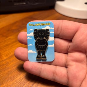 Pode incluir: Pin de esmalte preto com uma figura estilizada com olhos riscados e cabe&ccedil;a redonda, em um cart&atilde;o azul com o texto "FULLSYNCHROPINS". O pin tem detalhes dourados nos p&eacute;s.