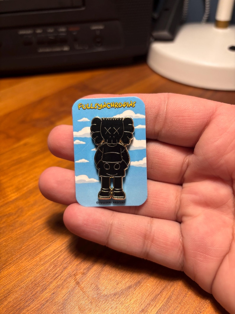 Pode incluir: Pin de esmalte preto com uma figura estilizada com olhos riscados e cabe&ccedil;a redonda, em um cart&atilde;o azul com o texto "FULLSYNCHROPINS". O pin tem detalhes dourados nos p&eacute;s.