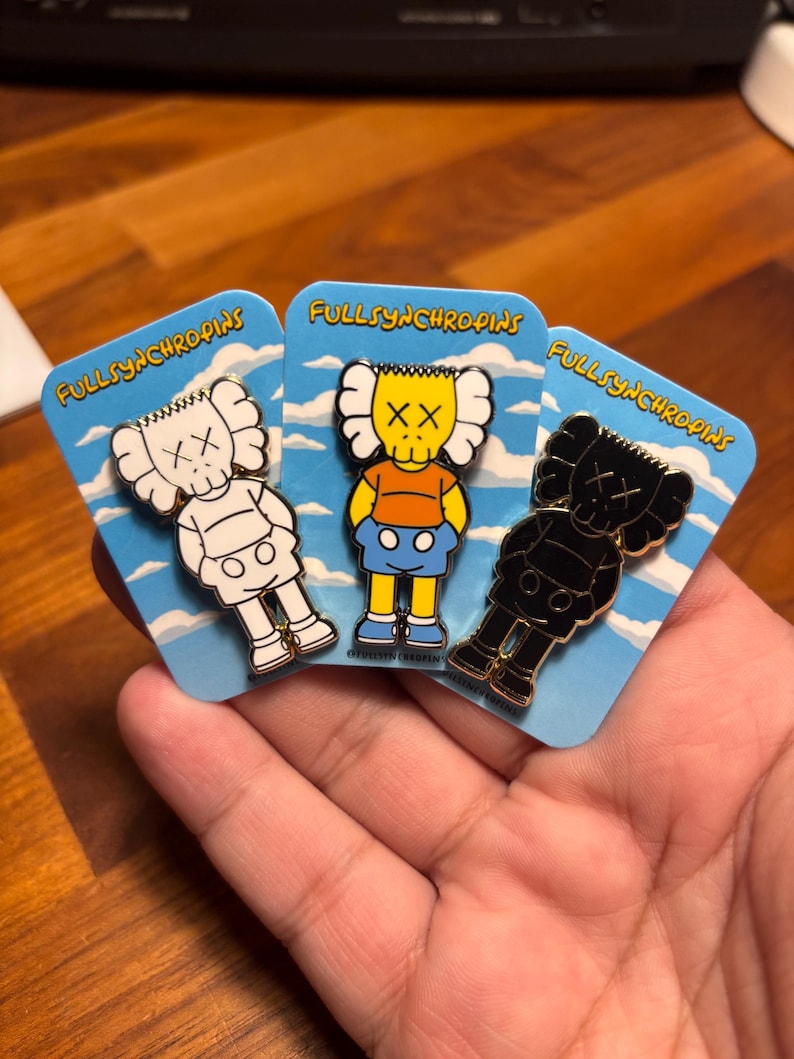 Pode incluir: Tr&ecirc;s pinos de esmalte com personagens de desenhos animados com olhos em forma de X. Um &eacute; branco, outro preto e outro amarelo com uma camisa laranja e cal&ccedil;&otilde;es azuis. Cada pino est&aacute; em um cart&atilde;o azul com o texto "FULLSYNCHROPINS".