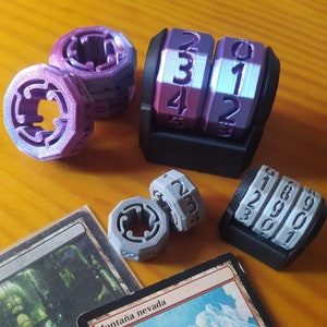 Puede incluir: Un conjunto de dados de seis caras con los números del 1 al 6 en cada cara. Los dados están hechos de plástico y son de diferentes colores. Hay dos conjuntos de dados, uno es morado y el otro es gris. Los dados están sobre una superficie de madera.