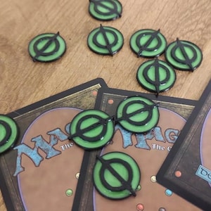 May include: Green and black plastic tokens with a symbol of a circle with a line through it. The tokens are on a wooden surface and a Magic: The Gathering card.