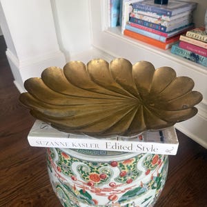 May include: A shallow, scalloped brass bowl with a radial, petal-like design. The bowl rests on a white book titled Suzanne Kasler Edited Style, which is placed atop a cylindrical ceramic garden stool featuring colorful painted figures and floral motifs.