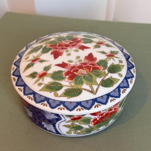 May include: A round, lidded ceramic trinket box. The box is white with a floral design featuring red flowers and green leaves. The rim has a blue and white pattern. The box is likely used for storing small items.