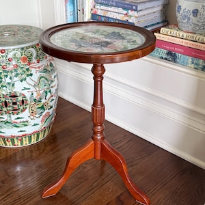 Op de afbeelding: Een kleine, ronde, houten bijzettafel met een donkerbruine afwerking. De tafel heeft een glazen blad met een bloemenontwerp eronder. Hij staat op een driepoot en is ongeveer 61 cm hoog.