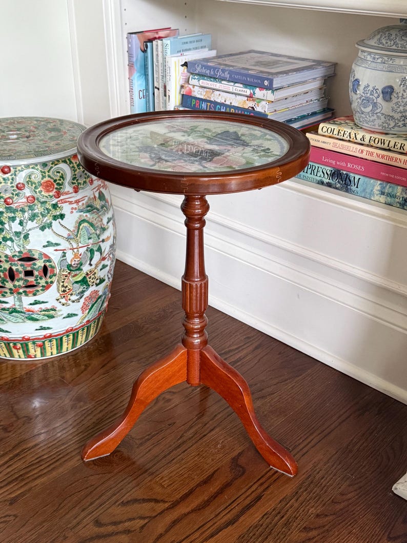 Op de afbeelding: Een kleine, ronde, houten bijzettafel met een donkerbruine afwerking. De tafel heeft een glazen blad met een bloemenontwerp eronder. Hij staat op een driepoot en is ongeveer 61 cm hoog.